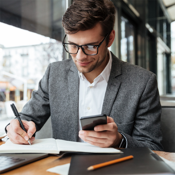 Empresário de blazer à mesa de trabalho, anotando num caderno com o celular na mão e o notebook ao lado.